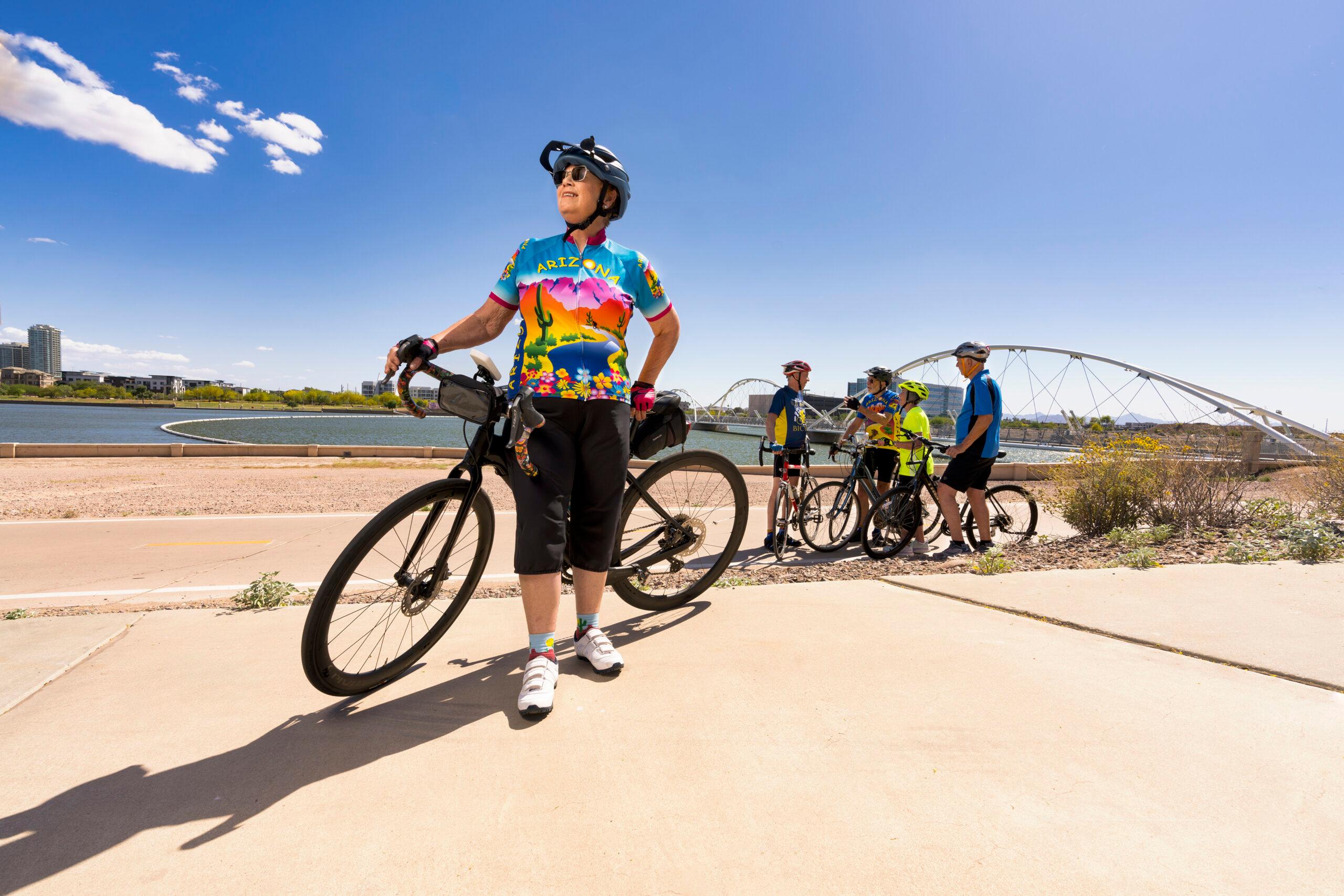 An older person stands with a bike in the foreground of the photo. A group of bikers stands in front of a picturesque lake in the background.