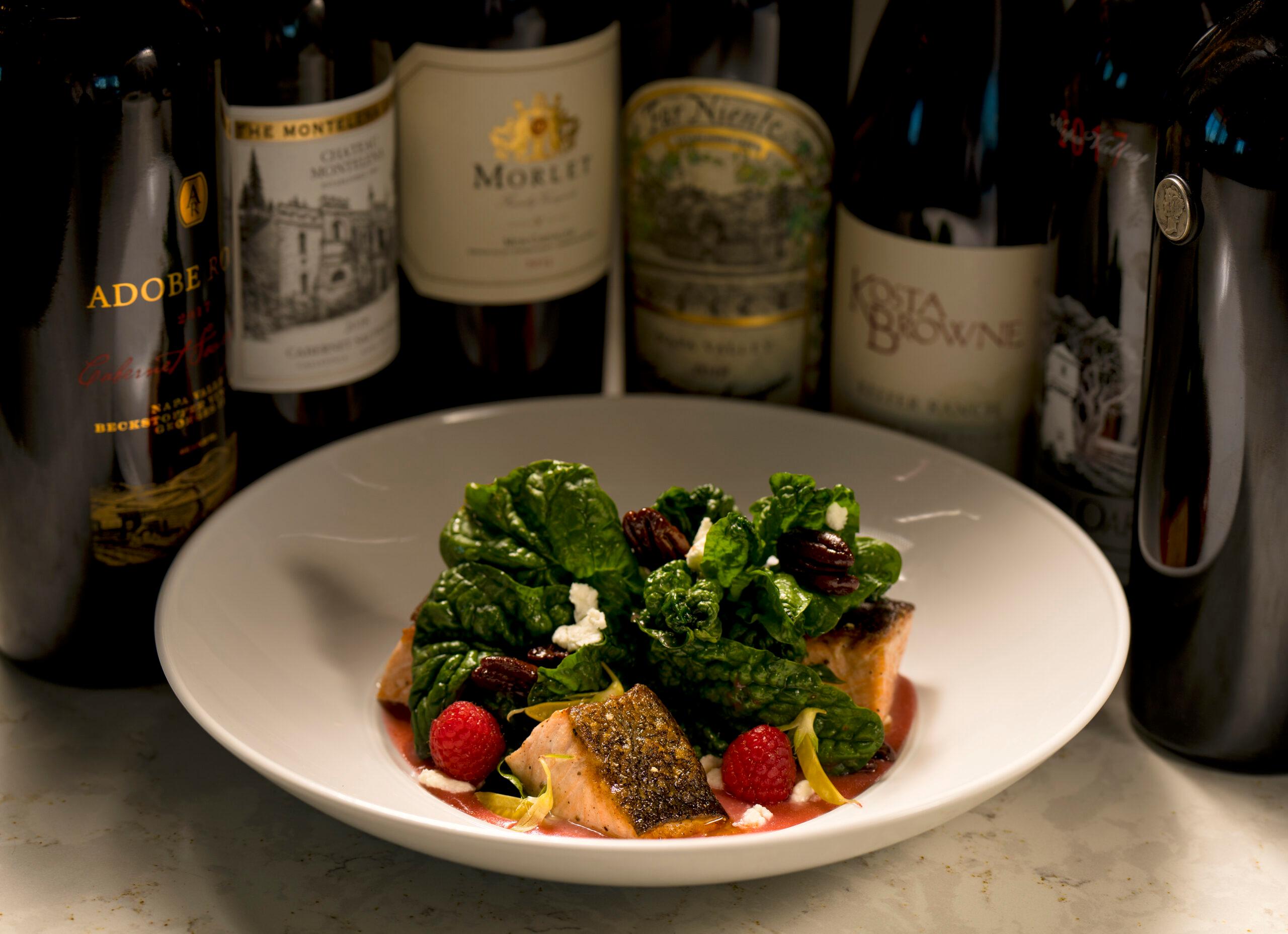 A gourmet plate of salmon and greens, with fancy bottles of wine in the background.