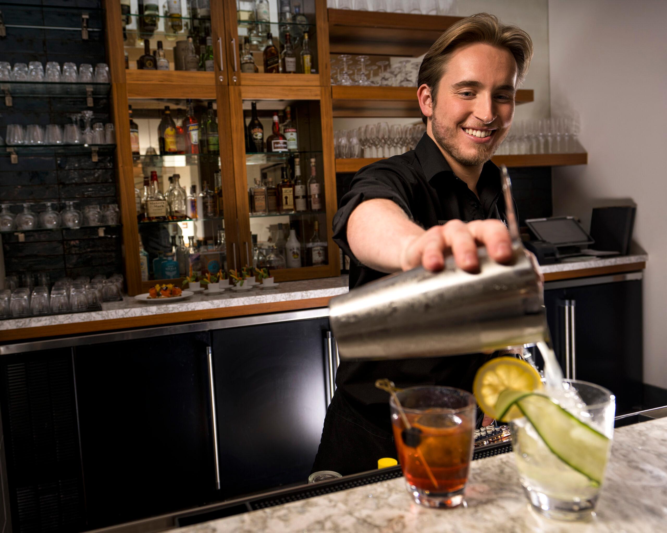 An expert mixologist pours a drink, garnished with lemon and cucumber.