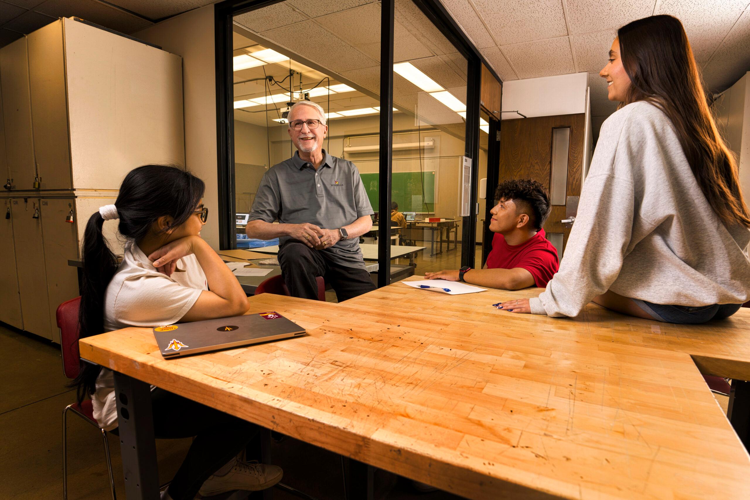 An older man teaches students at a table. The students look happy and engaged.