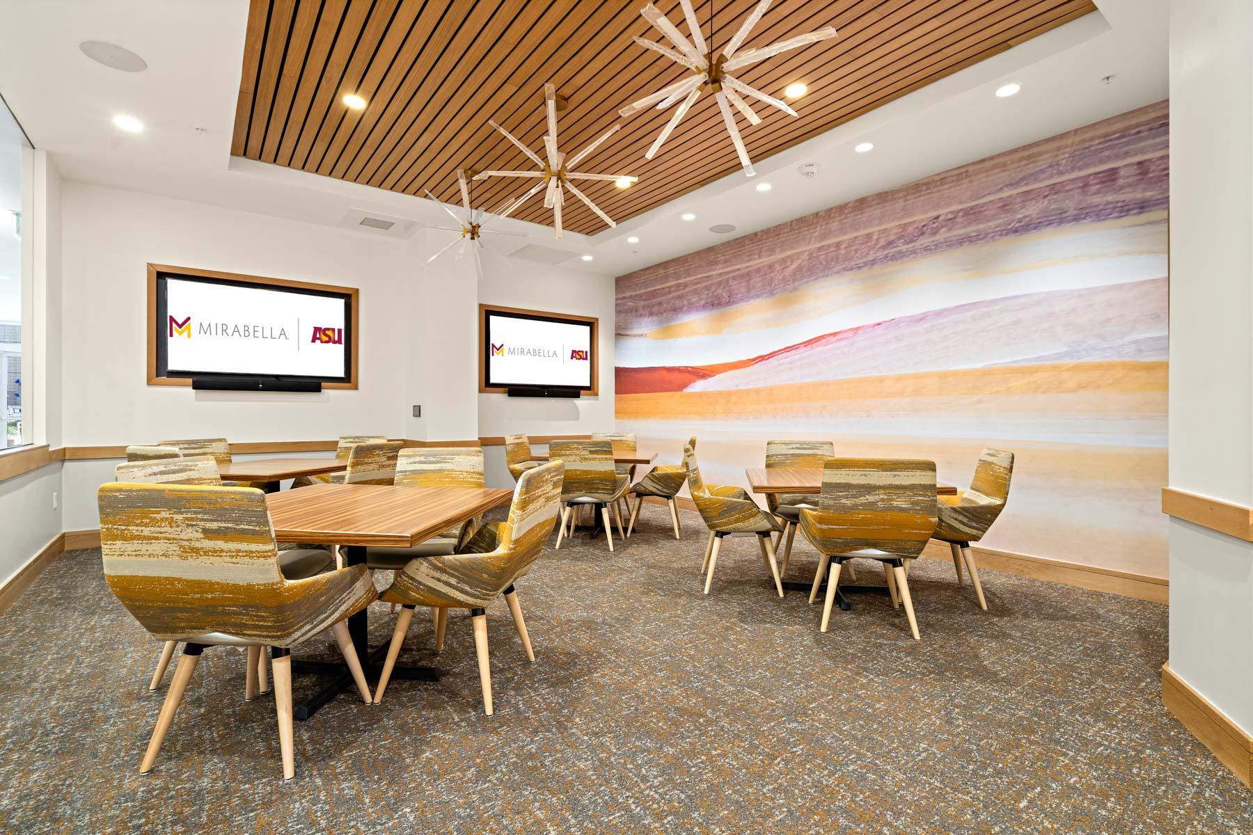 A room with tables and chairs as well as two TV screens.