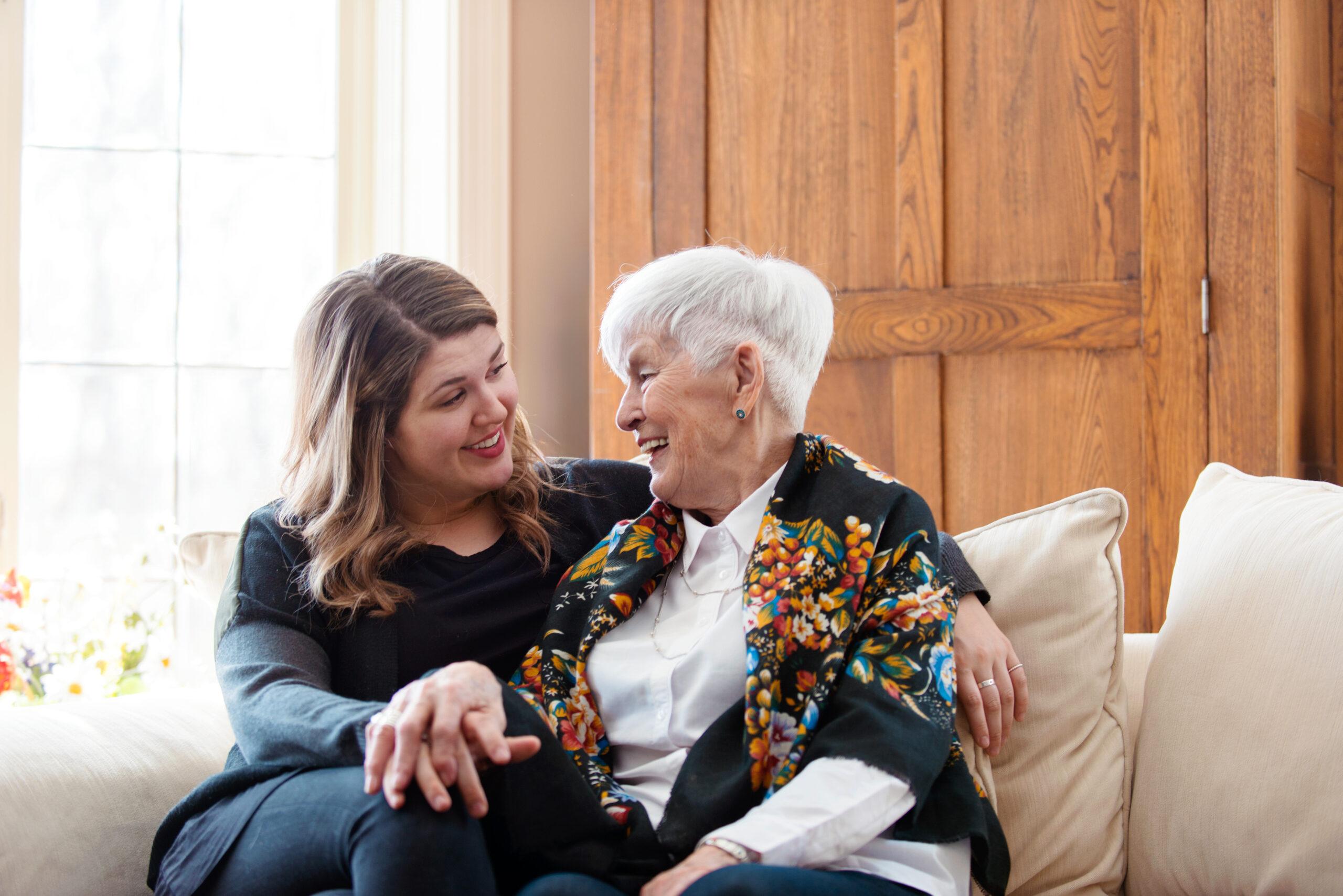 An adult woman sits on the couch with her mother. They are smiling and looking at each other.