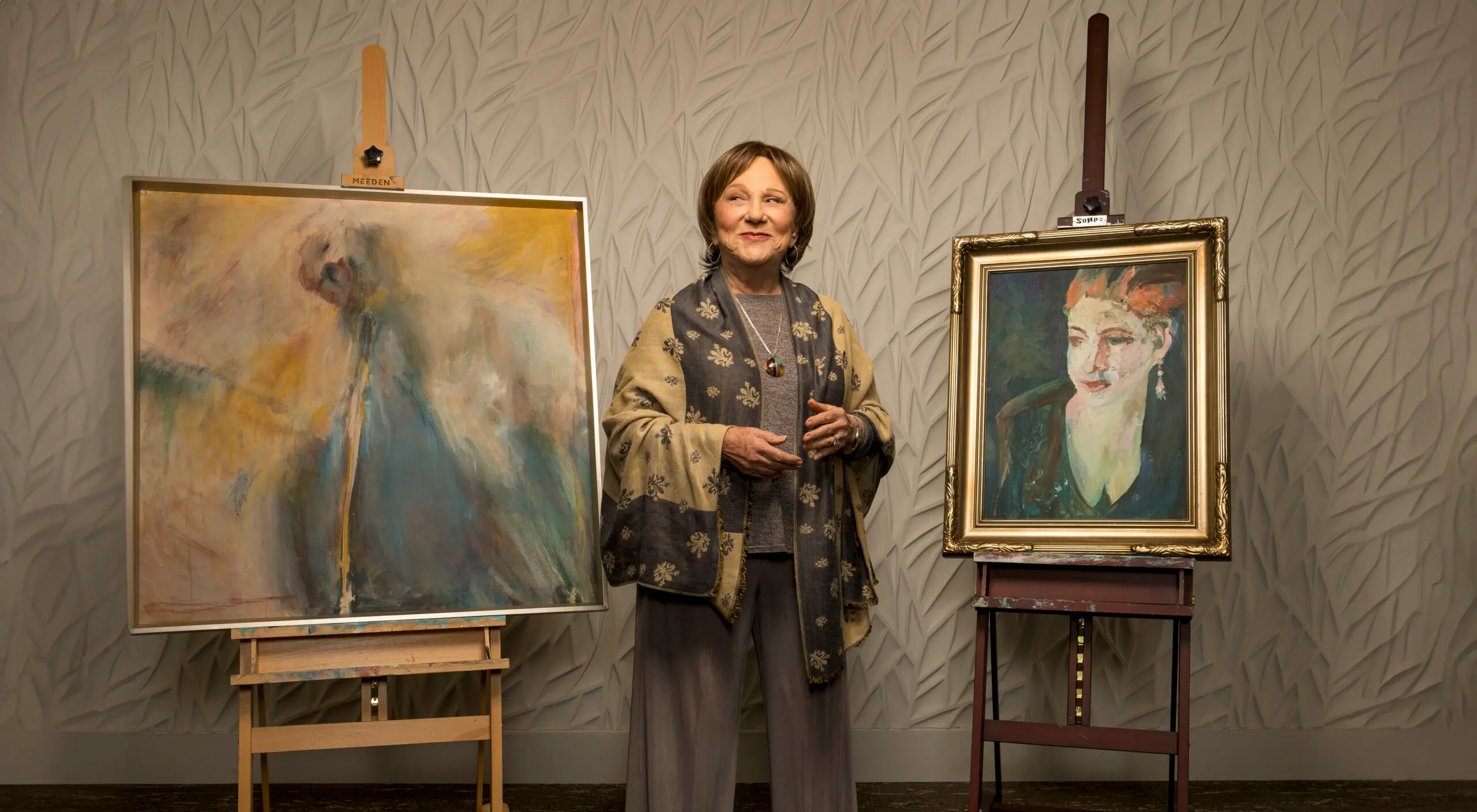 A woman stands with two paintings on easels with a proud expression.